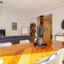 Salón comedor del departamento Nicasio en La Latina, Madrid, con mesa de madera brillante, sofá gris, televisión, espejo decorativo y detalles modernos, gestionado por Stag Properties y destinado a alquiler vacacional